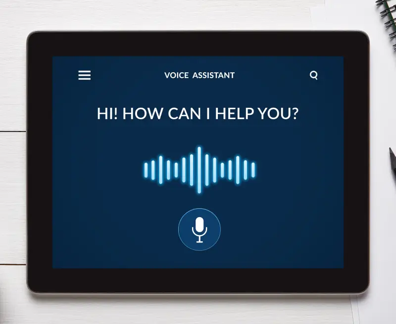 A concept of a voice assistant on a tablet sitting with the text Hi! How can I help you? on the screen.